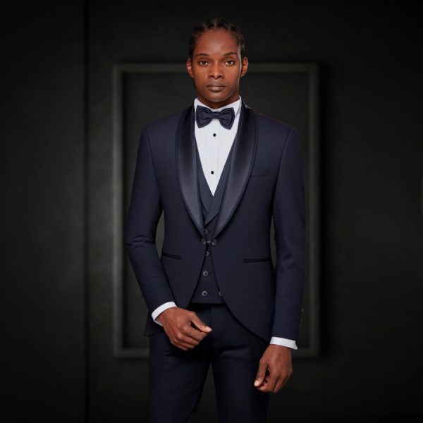 A man in a navy blue tuxedo and tie standing in front of a white background.